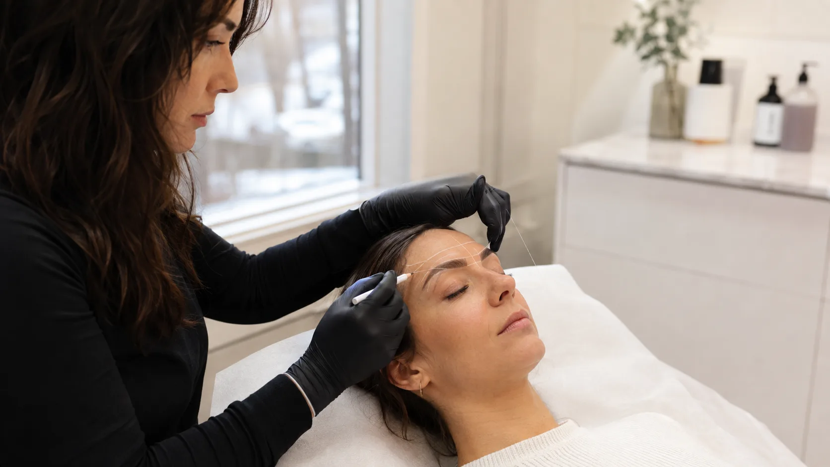 Cosmetic tattoo artist mapping a client’s brows inside a winter-lit studio in Canada