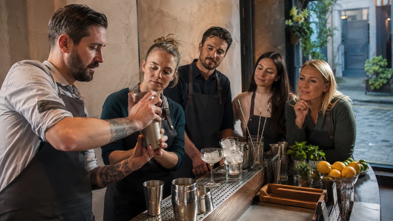 A tutor showing shaker technique at a rendered-wall bar in Ireland, a wet courtyard through the door