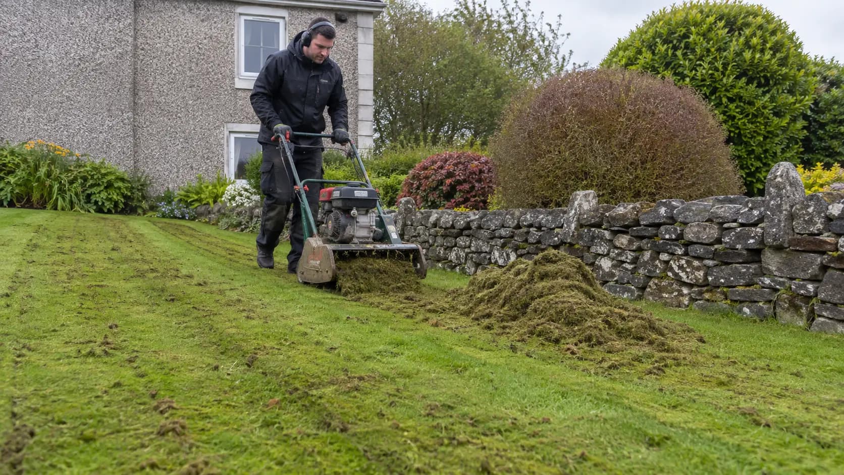 An operator scarifies a lawn beside a dry stone wall at an Irish house