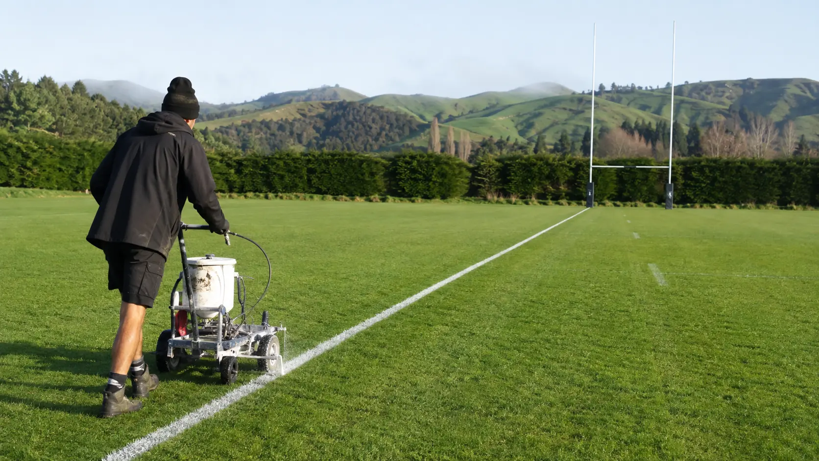 An operator walks a line marker across a New Zealand rugby field below green hills