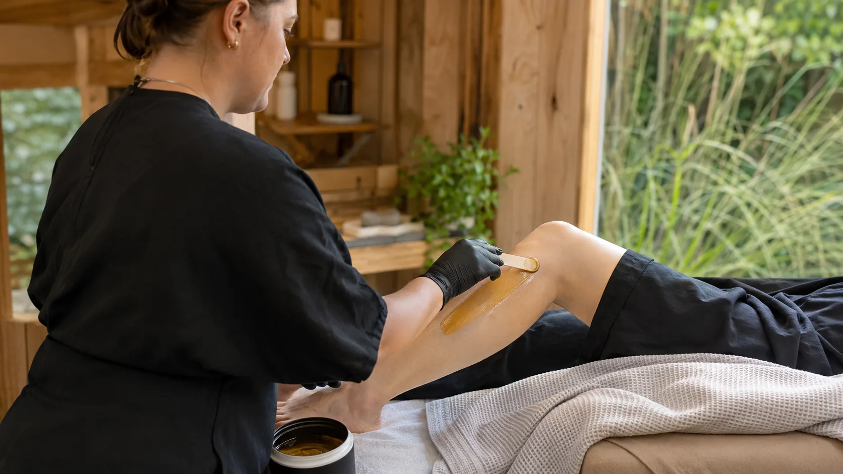 Waxing therapist applying leg wax for a client inside a timber-lined studio in New Zealand